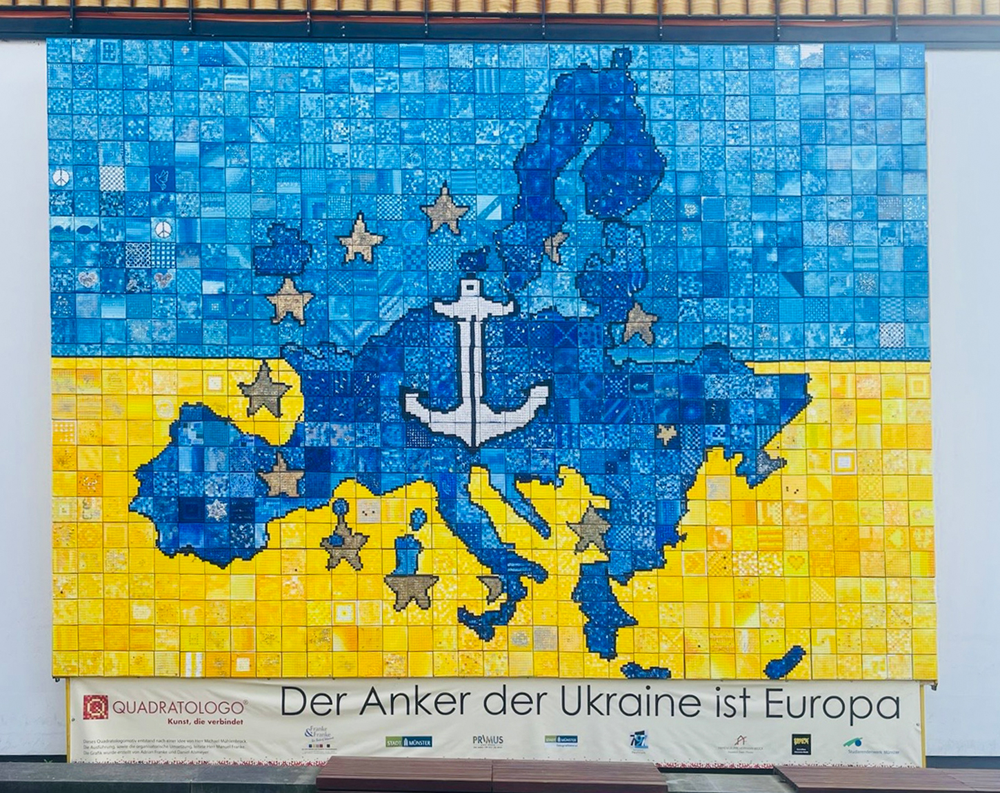 Das Foto zeigt das Wandbild, welches an der Wohnanlage Bismarckallee steht. Auf dem Bild ist ist Europa mit einem silbernen Anker zu sehen, um den Anker sind silberne Sterne Das Bild ist in gelb und blau unterteilt.